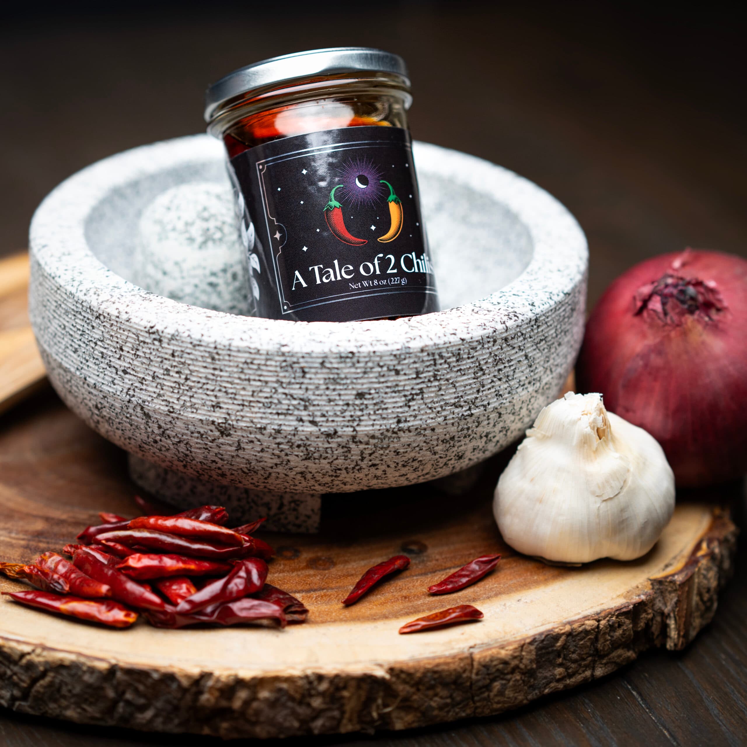 A jar of chili oil in a stone mortar surrounded by dried chilis, garlic, and red onion